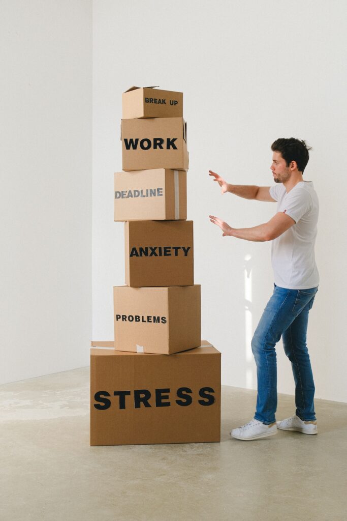 A tall stack of cardboard boxes threatens to overwhelm a person standing next to it. The person braces with arms outstretched, expecting the boxes to fall down. The boxes are labelled and say from top to bottom: Break up, work, deadline, anxiety, problems, stress.
